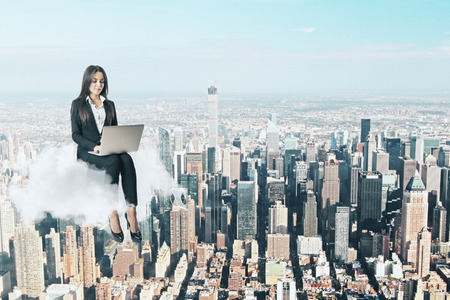Attractive young businesswoman using laptop while sitting on cloud. City background. Work freedom and freelance conceptの写真素材