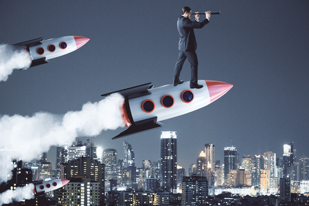 Businessman using telescope while standing on launching rocket. City background. Future and startup conceptの写真素材