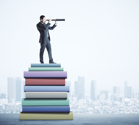 Attractive young european businessman using telescope while standing on pile of books on blurry city background. Education and vision concept.の写真素材