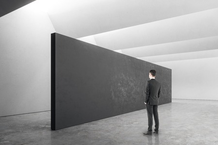 Thoughtful young businessman standing in modern exhibition hall interior with copy space on wall....
