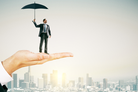 Big hand holding young businessman with umbrella on bright city background with sunlight. Protection and success conceptの写真素材