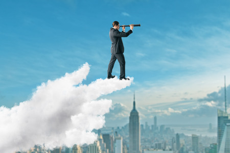 Young businessman using telescope while standing on pointing cloud hand on New York city background. Vision and success conceptの写真素材
