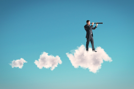 Young businessman using telescope while standing on cloud on sky background. Vision and success conceptの写真素材