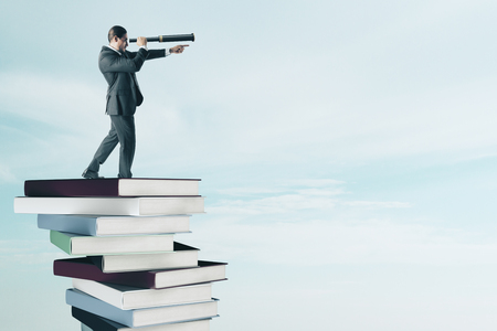 Side view of young businessman with binoculars standing on pile of books. Vision and education conceptの写真素材