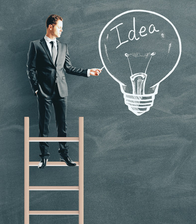 Young businessman standing on ladder while drawing idea lamp on chalkboard wall background. Innovation and power conceptの写真素材