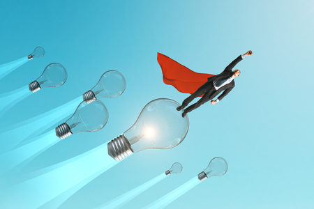 Young superhero businessman with cap flying with lamps on blue sky background. Leadership and innovation concept.の写真素材