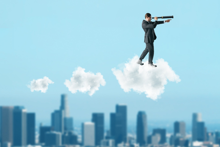 Young businessman using telescope while standing on cloud on blurry sky background. Vision and spy conceptの写真素材