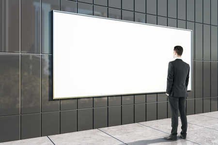 Young businessman looking at empty black tile wall with billboard and city reflections. Mock up,の写真素材