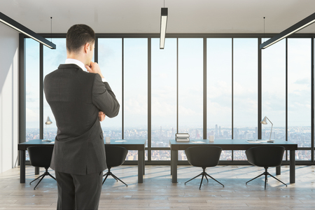 Thoughtful young businessman standing in modern office with panoramic city viewの写真素材