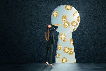 Businesswoman looking at bitcoins through keyhole opening in wall. Cryptocurrency and commerce conceptの写真素材