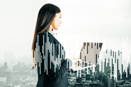 Attractive young businesswoman using laptop on abstract city background with forex chart. Trade and communication concept. Double exposureの写真素材