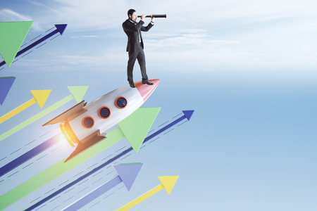 Side view of young businessman with telescope standing on launchging rocket on sky background with colorful arrows. Startup, growth and up concept.の写真素材