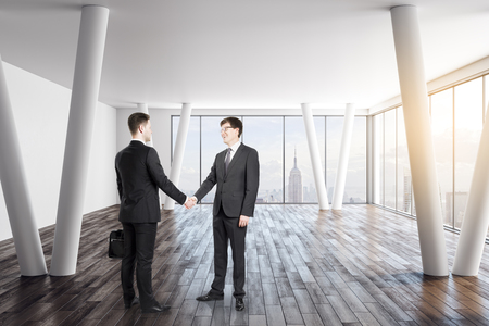 Attractive european businessmen shaking hands in modern office interior with city view and daylight. Teamwork and partnership concept.の写真素材