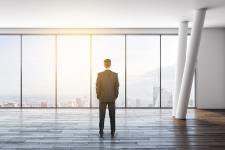 Back view of young businessman in modern empty white office interior with wooden floor and panoramic New York city view. Research concept.の写真素材