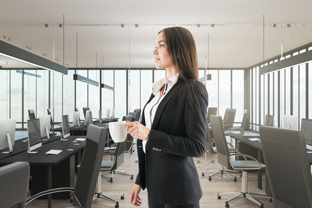 Pretty young european businesswoman drinking coffee in modern office interior. Breakfast and morning concept.の写真素材