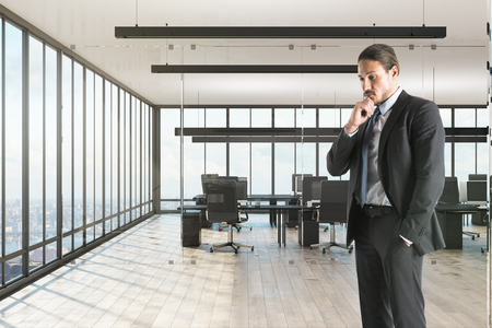 Thinking young businessman standing in modern office with panoramic city view.の写真素材