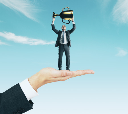 Hand holding businessman with golden cup on sky background. Leader and success conceptの写真素材