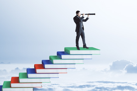 Businessman with binoculars standing on top of book pile ladder. Sky background. Education and vision conceptの写真素材