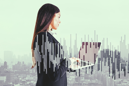 Attractive young businesswoman using laptop on abstract city background with forex chart. Network and communication concept. Double exposureの写真素材
