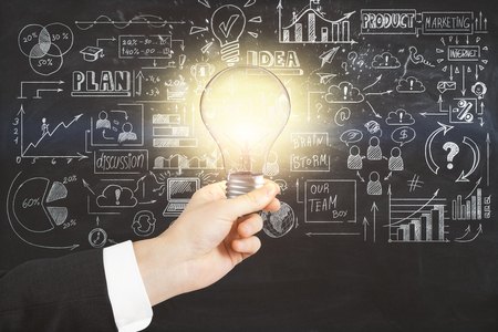 Close up of businessman hand holding glowing lamp on concrete wall background with creative business sketch. Idea and finance conceptの写真素材