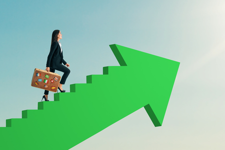 Young businesswoman wth luggage climbing abstract green arrow ladder on sky background. Career promotion and travel concept.の写真素材