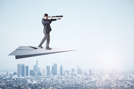 Young businessman using telescope to look into the distance while standing on paper plane on sky and city background. Research and direction conceptの写真素材
