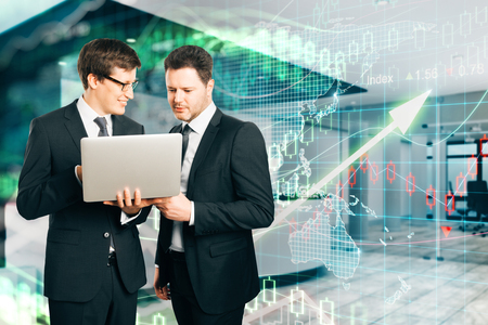 Two young businessmen using laptop together in modern office interior with increasing forex chart arrow. Teamwork and investment concept. Double exposureの写真素材