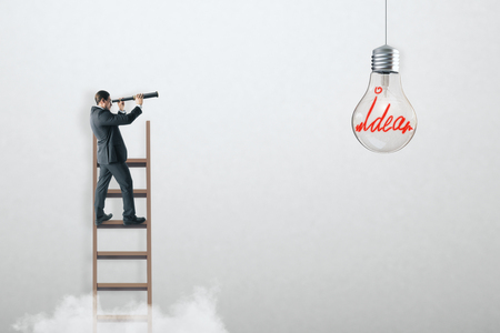 Businessman on ladder using telescope to look at hanging light bulb. White concrete wall background. Idea and achievement conceptの写真素材