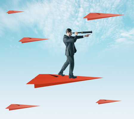 Young businessman using telescope to look into the distance while standing on red paper plane on sky background. Research and forecast conceptの写真素材