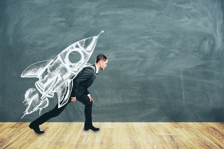 Young businessman carrying creative rocket sketch on his back on chalkboard wall background. Startup and work conceptの写真素材