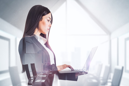 Side view of attractive young businesswoman using laptop in blurry office interior. Technology and communication concept. Double exposureの写真素材