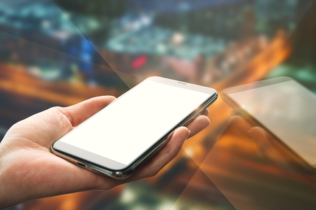 Hand holding empty white smartphone with reflection on blurry night city background. Mock upの写真素材