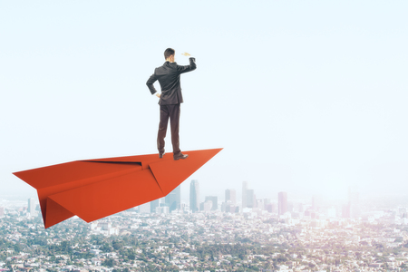 Young businessman looking into the distance while standing on red paper plane. Sky and city background. Freedom and vision conceptの写真素材