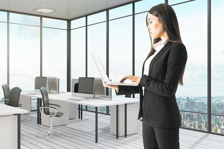 Attractive young european businesswoman using laptop in modern white office interior. Technology and communication concept.の写真素材