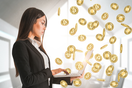 Side view of attractive young businesswoman using laptop in abstract blurry office interior with bitcoin rain. Technology and cryptocureency concept.の写真素材