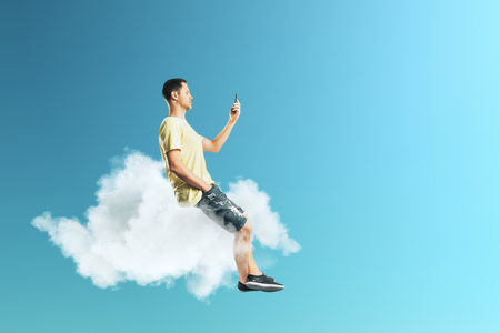 Side view of young european guy on cloud using smartphone. Sky background. Freedom and technology conceptの写真素材