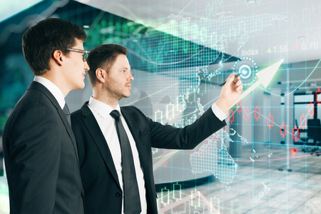 Two young businessmen standing in modern office interior with forex chart. Teamwork and finance concept. Double exposureの写真素材