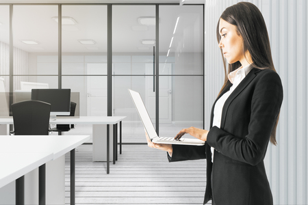 Pretty young european businesswoman using laptop in modern white office interior. Technology and communication concept.の写真素材