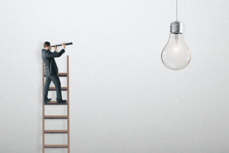 Businessman on ladder using telescope to look at hanging light bulb. White concrete wall background. Idea and solution conceptの写真素材