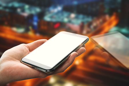 Hand holding empty white mobile phone with reflection on blurry night city background. Mock upの写真素材