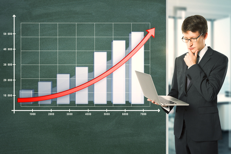 Attractive young businessman with laptop standing next to blackboard with business chart and arrow. Financial growth and presentation conceptの写真素材