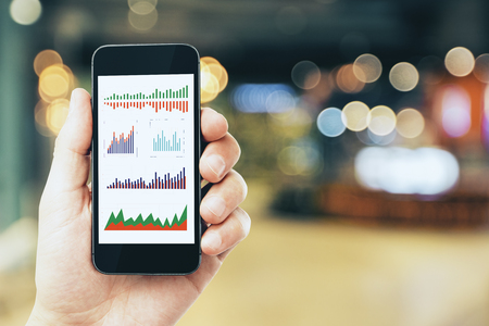 Businessman hand holding cellphone with business charts on blurry bokeh interior background. Device and mobile finance conceptの写真素材