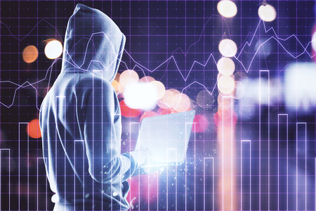 Hacker in hoodie holding laptop and standing on abstract blurry night city background with forex chart grid. Investment and hacking concept. Double exposureの写真素材