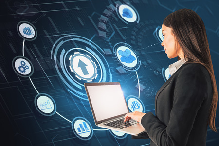Side view of attractive young businesswoman using laptop on blue background with abstract business interface. Technology and finance concept.の写真素材