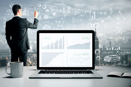 Young businessman drawing creative business sketch on abstract city background with charts on laptop screen and coffee cup. Report and finance conceptの写真素材