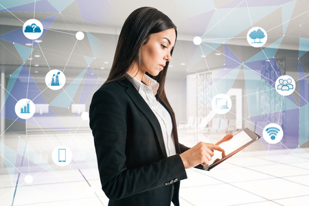 Side view of attractive young businesswoman using tablet on blurry office interior background with abstract business interface. Technology and network concept. Double exposureの写真素材