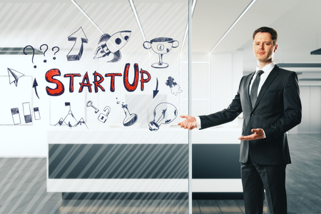 Attractive young businessman standing in office reception interior with startup sketch on glass.  Start up and success concept.の写真素材