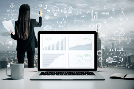 Back view of young businesswoman drawing creative business sketch on abstract city background with charts on laptop screen and coffee cup. Report and workshop conceptの写真素材