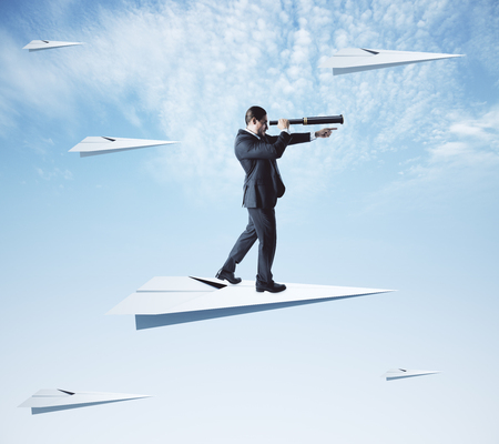 Young businessman using telescope to look into the distance while standing on paper plane on sky background. Research and leadership conceptの写真素材
