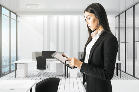 Pretty young european businesswoman using tablet in modern white office interior. Technology and communication concept.の写真素材